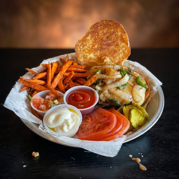 A burger with grilled onions and peppers, sweet potato fries, and sides on a metal plate.